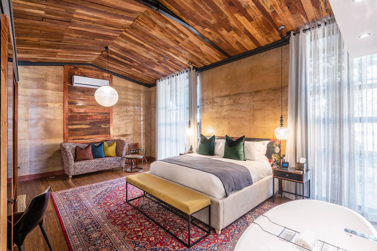 A bedroom with a wooden ceiling, a large bed with multiple pillows, a bench, a bedside table with a lamp, and a large window with curtains.