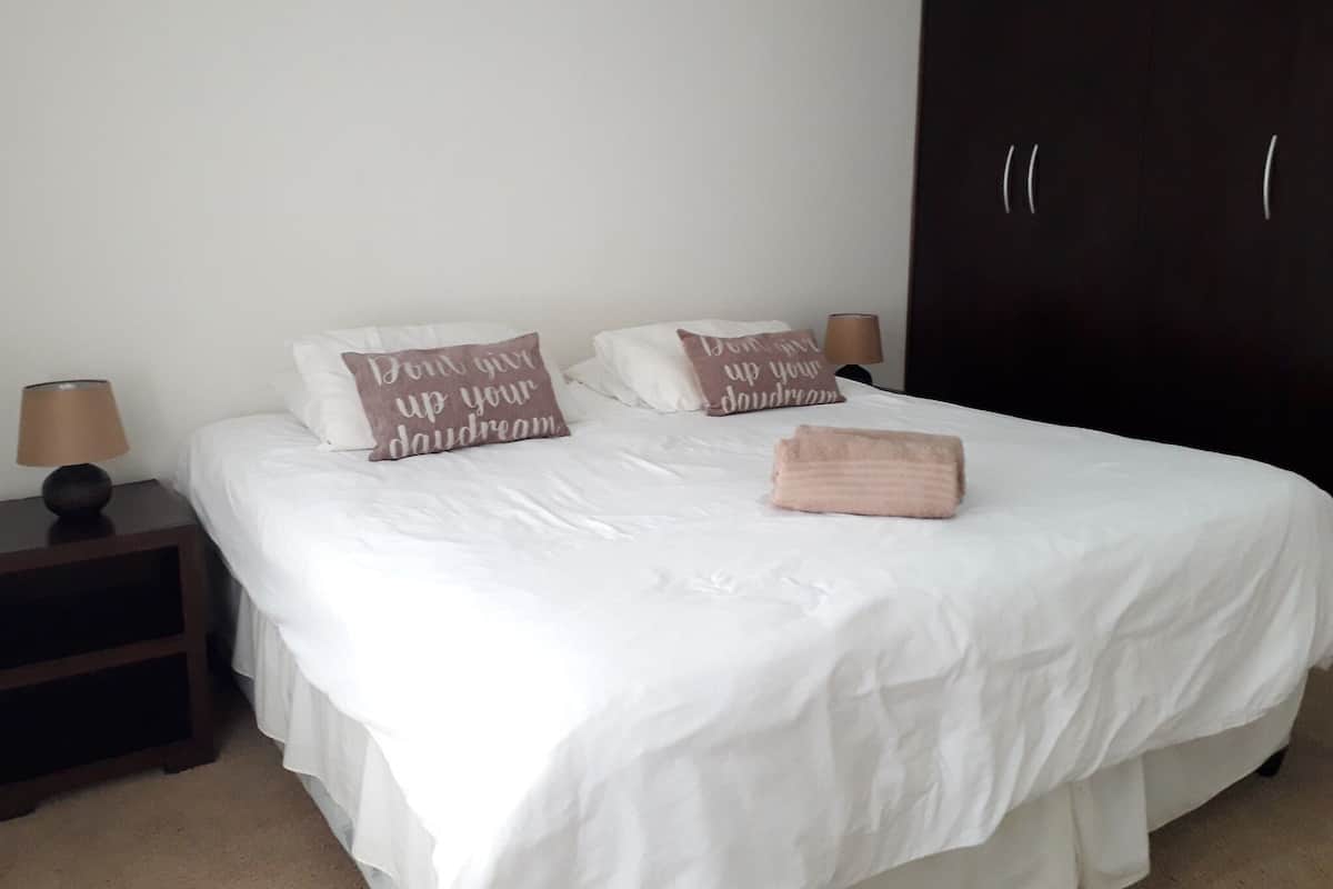 Una cama bien hecha con ropa blanca y dos almohadones decorativos, un armario de madera oscura y una mesita de noche con una lámpara.