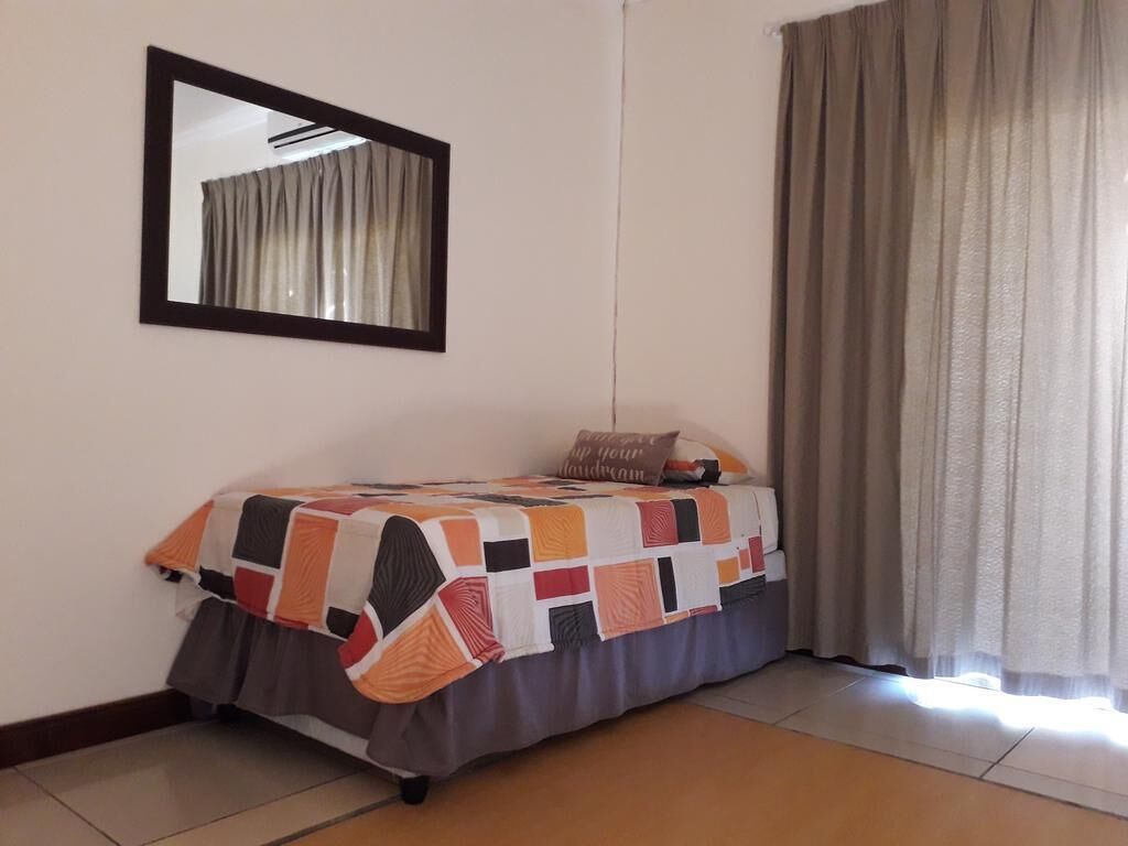 A single bed with a colorful quilt, a mirror on the wall, and a window with curtains.