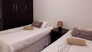 Ein Zimmer mit zwei Betten, einem Schrank, einem Nachttisch mit Lampe und dekorativen Kissen.