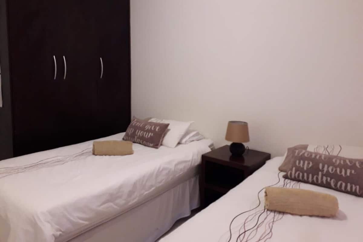 Una habitación con dos camas, un armario, una mesita de noche con lámpara y almohadas decorativas.