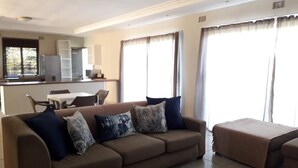 Ein Wohnzimmer mit einer Couch, einem Hocker und einem Essbereich im Hintergrund.