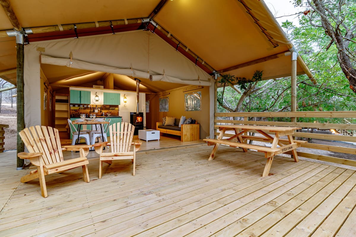 Eine Holzterrasse mit Adirondack-Stühlen und Picknicktischen, ein Küchenbereich mit Schränken und Herd sowie Blick auf Bäume durch das Blätterdach.