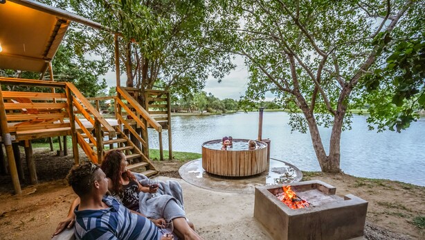 Um casal relaxando em um banco perto de um deck de madeira com vista para um lago, com uma lareira e uma banheira de hidromassagem por perto.