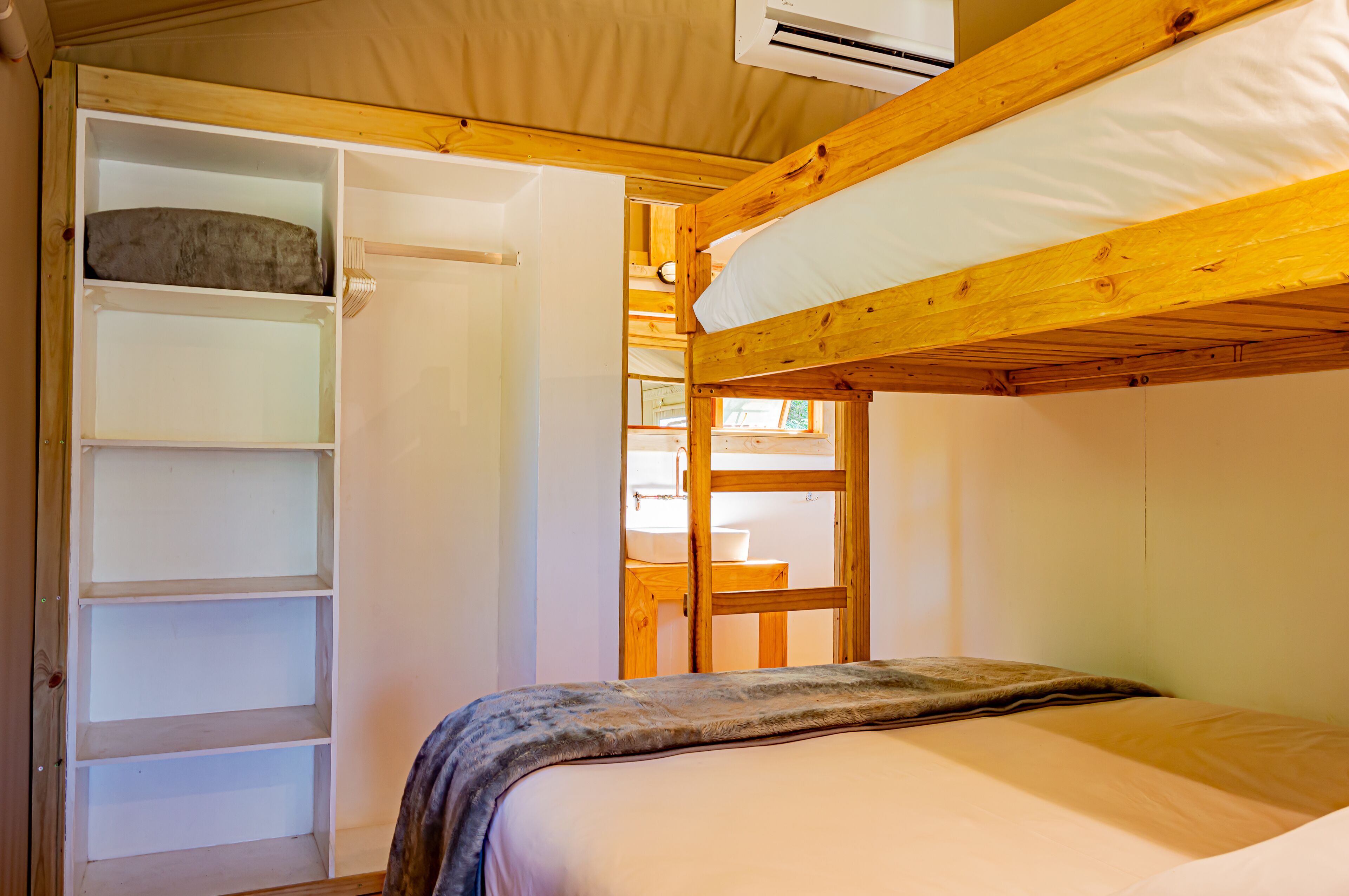 A bunk bed with a wooden headboard, a white mattress, and a blanket. There is a wooden shelf unit with multiple compartments and a white door.