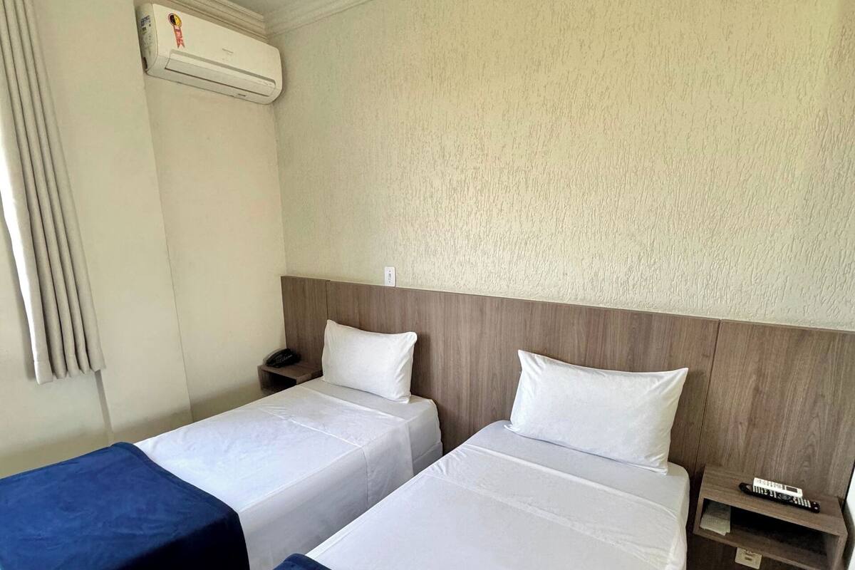Hotellihuone, jossa on kaksi sänkyä, yöpöytä, jossa on kaukosäädin, ja seinälle asennettu ilmastointilaite.