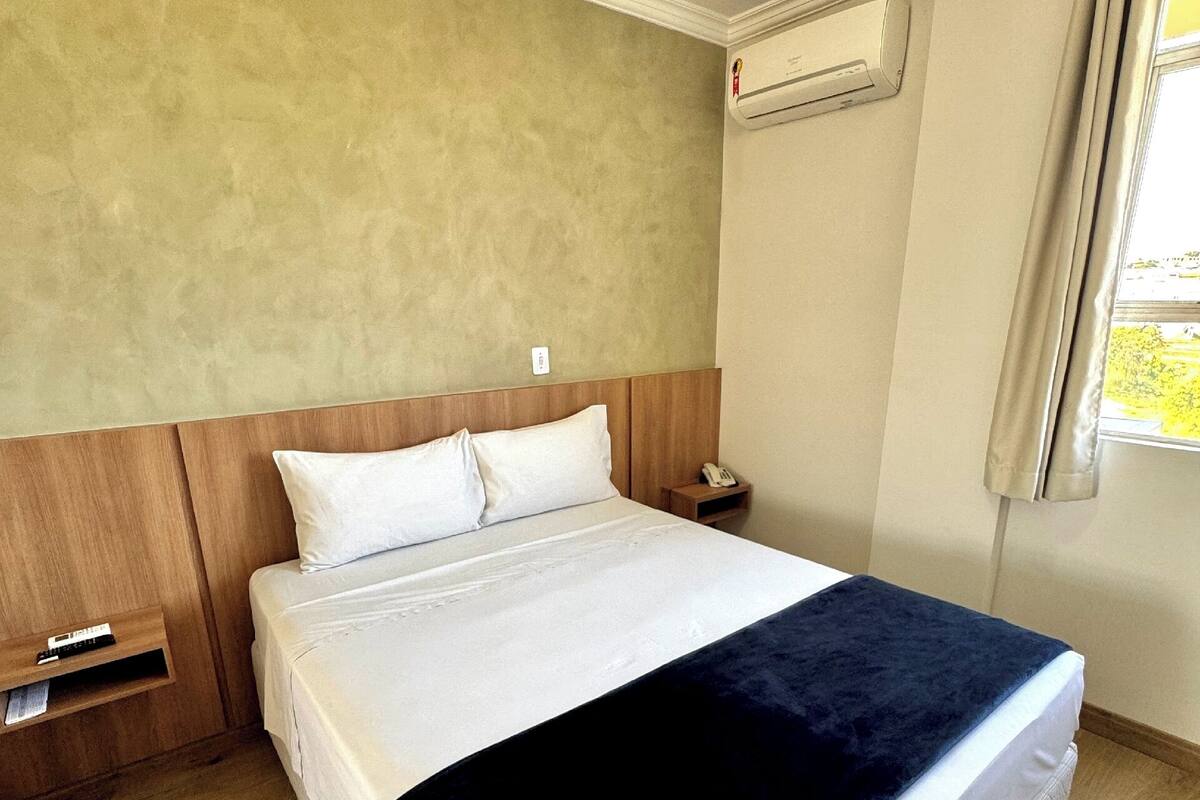 Hotellihuone, jossa on puinen sängynpääty, valkoinen päiväpeite, tummansininen viltti, yöpöytä, jossa on puhelin, ja seinälle asennettu ilmastointilaite.