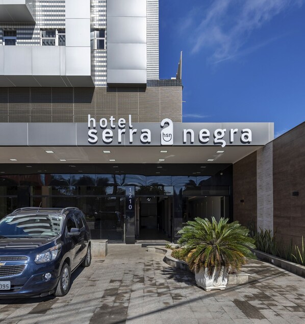The entrance to Hotel Serra Negra with a modern design, a car parked in front, and a large potted plant beside the entrance. - Hotel Serra Negra (Betim)