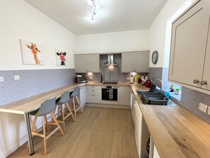 A compact kitchen with a wooden countertop, a built-in oven, a microwave, and a sink. There are two bar stools and a small window with plants.