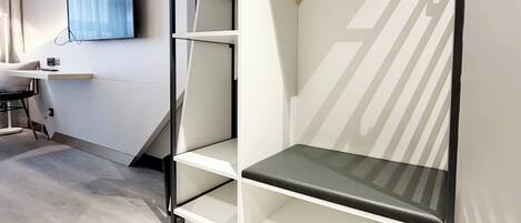 Una habitación moderna con pared blanca, televisor montado, escritorio y estantería empotrada con un asiento negro.