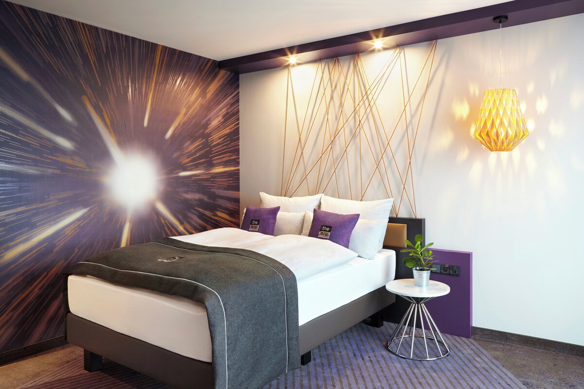 A modern hotel room with a large bed, a bedside table with a plant, and a unique wall design.