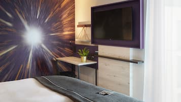 A modern hotel room with a bed, a mounted TV, a small desk with a plant, and a wall with a starburst pattern.