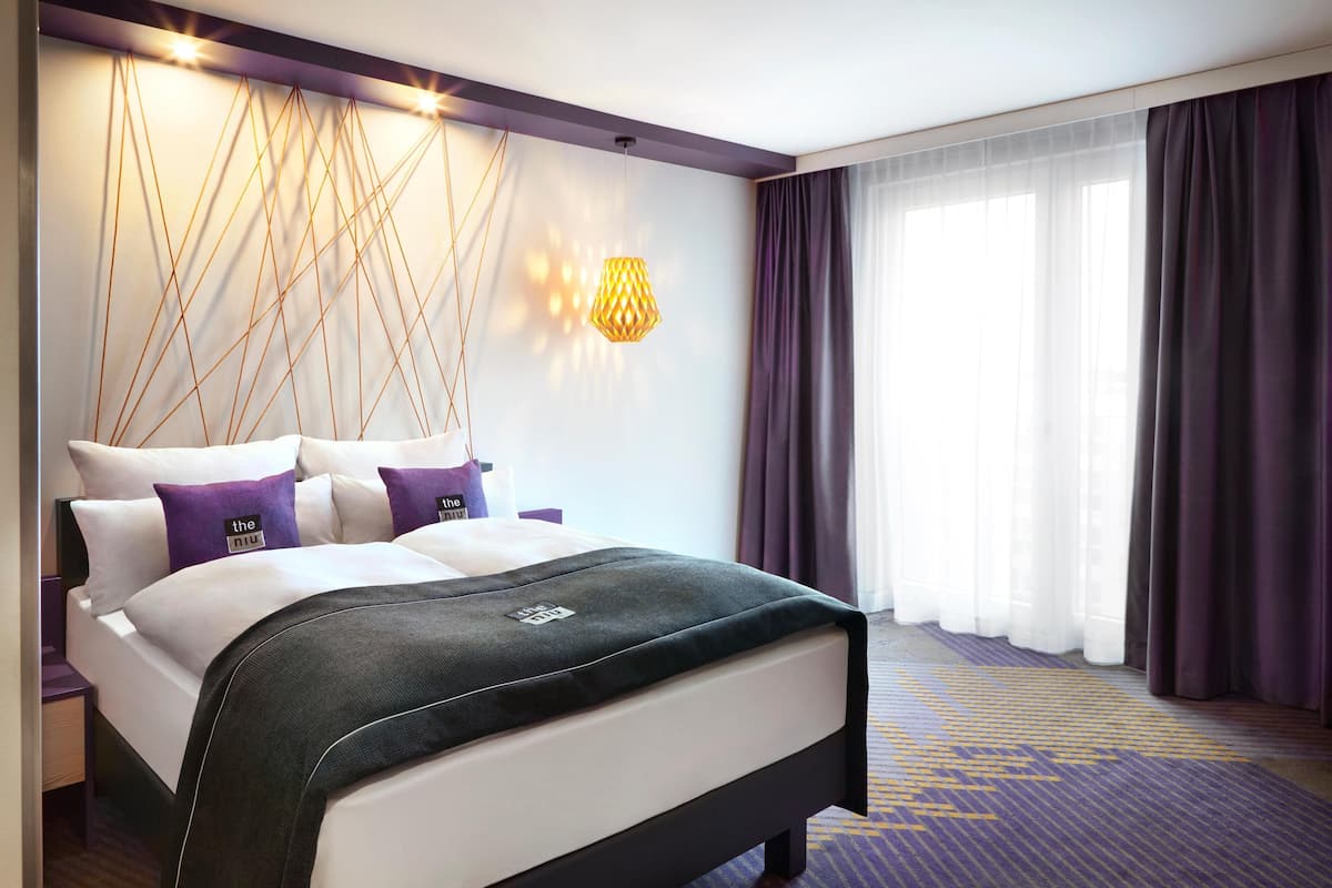 A hotel room with a bed, a wall-mounted lamp, a window with curtains, and a patterned carpet.