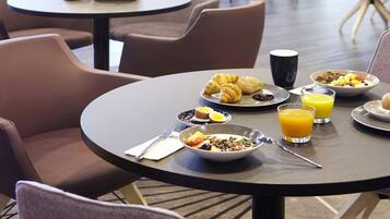 Une table dressée pour le petit-déjeuner, avec des tables rondes, des chaises et une variété d’aliments, notamment des croissants, des œufs et du jus d’orange.