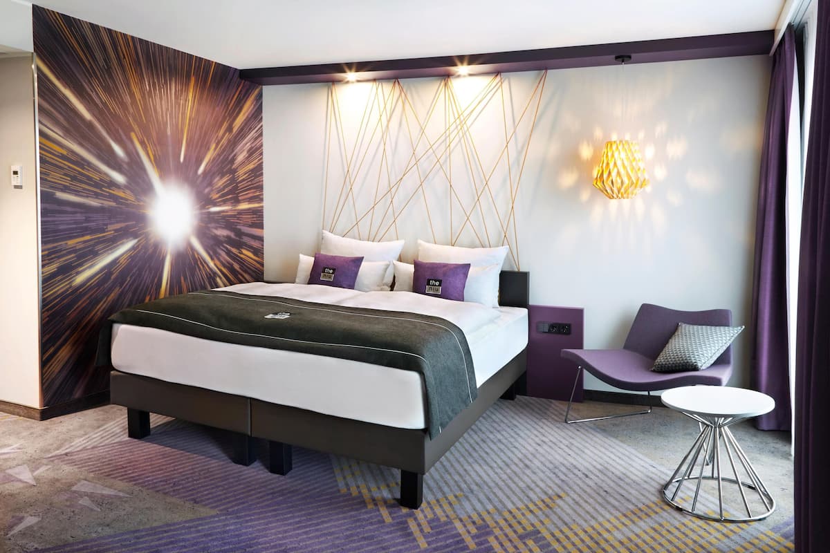 A modern hotel room with a large bed, a small table, and a chair. There is a wall with a starburst design and a wall-mounted light fixture.