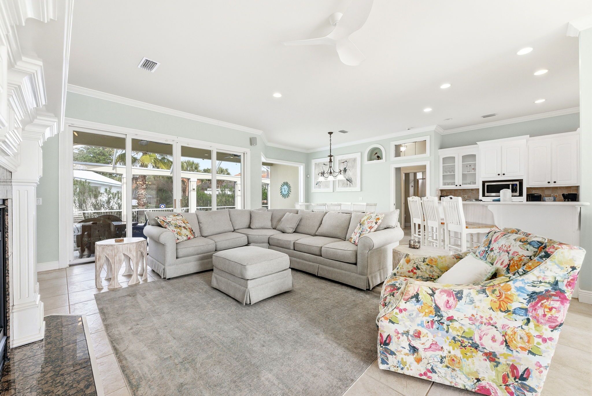 Een ruime woonkamer met een modulaire bank, een fauteuil met bloemenprint en een eethoek met witte kasten en een keuken op de achtergrond.