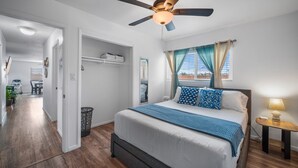 A bedroom with a bed, a ceiling fan, a window with curtains, a bedside table with a lamp, and a hallway leading to a bathroom. - Family Retreat w/ Theater, Pool & Outdoor Dining (Chula Vista)
