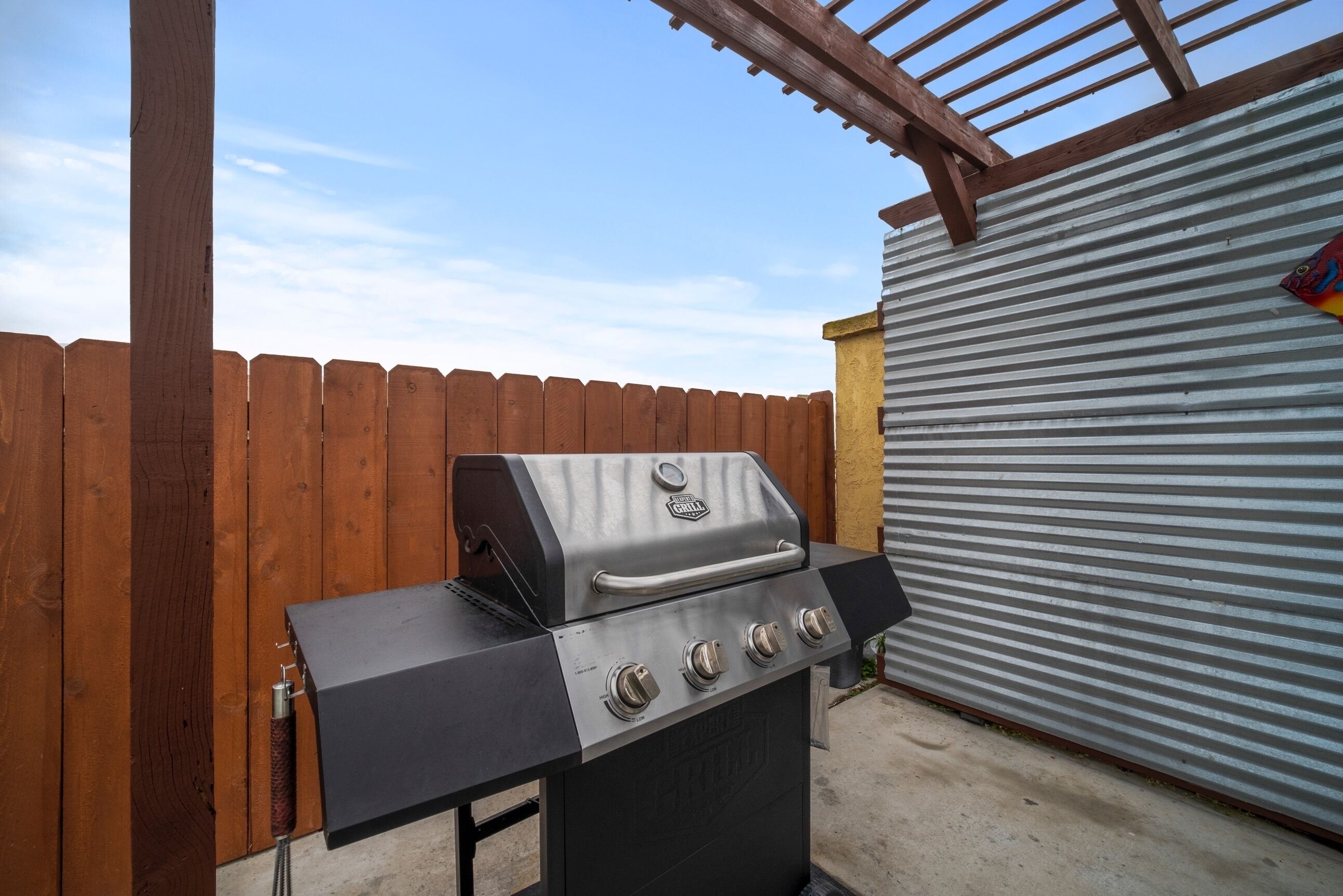 A charcoal grill with four burners and a side burner, positioned under a wooden pergola.
