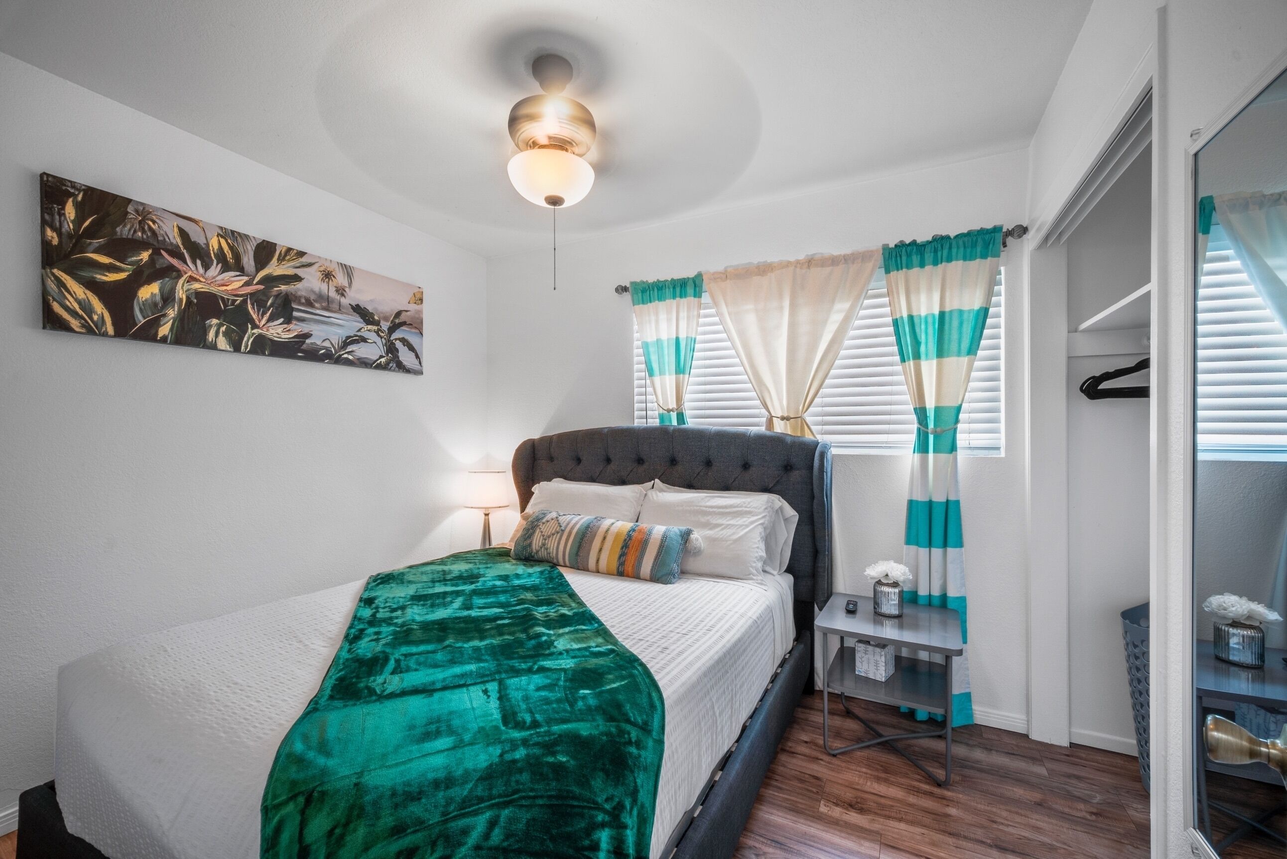Une chambre à coucher comprenant un lit, une table de chevet, un ventilateur de plafond, une fenêtre avec des rideaux et un tableau accroché au mur.