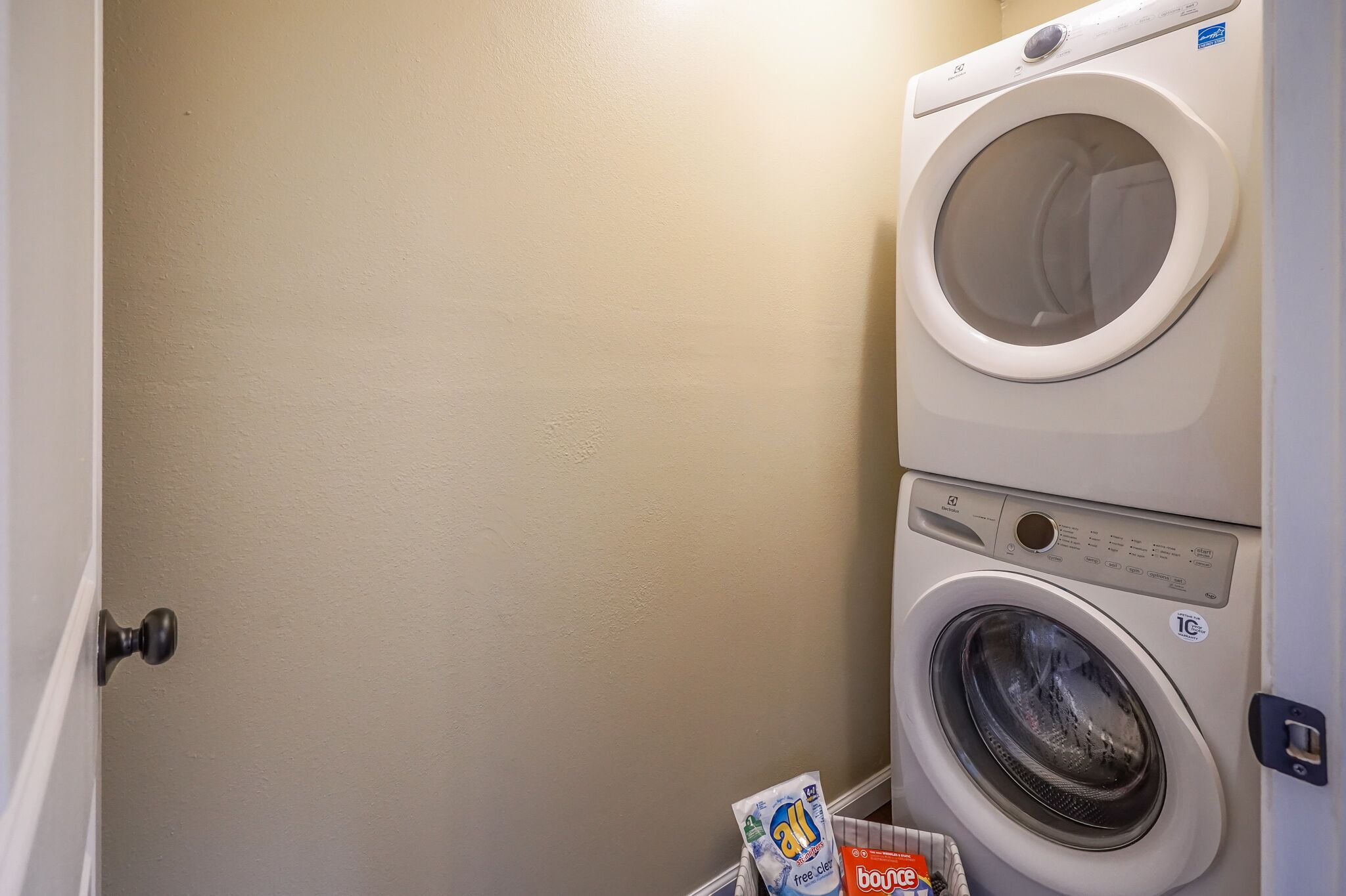 A laundry room with a front-loading washer and dryer, a door with a round doorknob, and a basket containing laundry detergent.