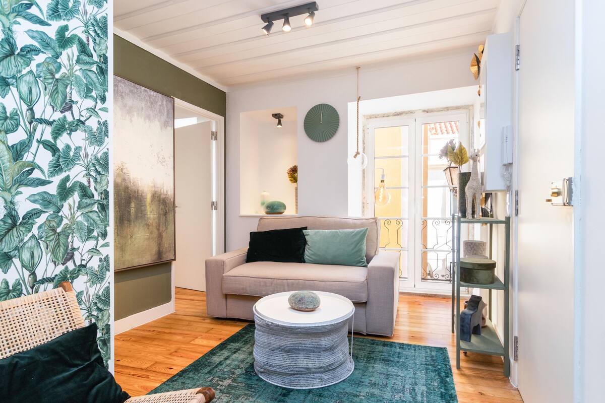 A living room with a beige sofa, a round coffee table, a green rug, and a wall with a tropical wallpaper.