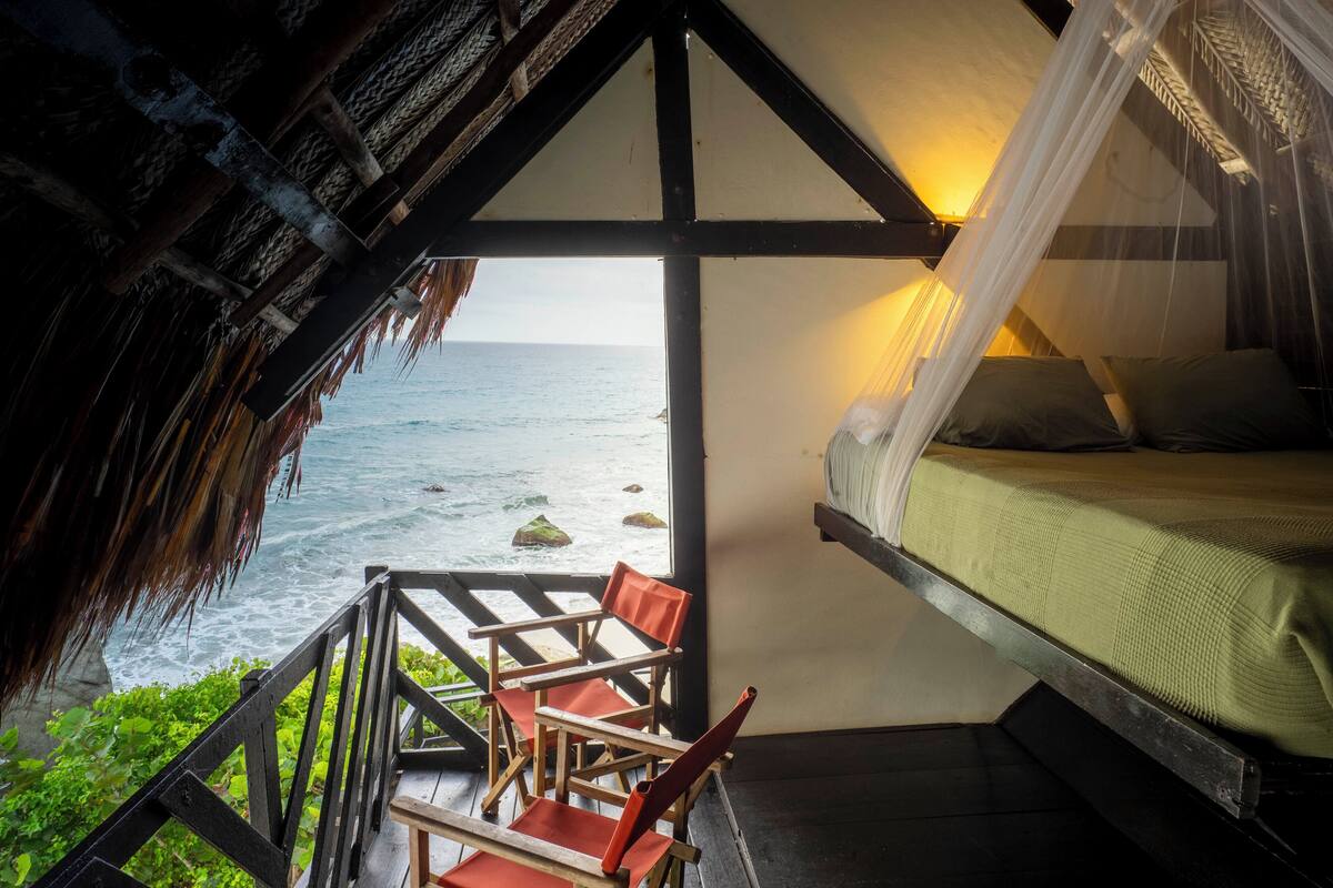 A bedroom with a bed, a view of the ocean, and a balcony with chairs.