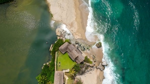 Vista aérea de una propiedad frente a la playa, con una estructura de techo de paja, piscina y abundante vegetación.