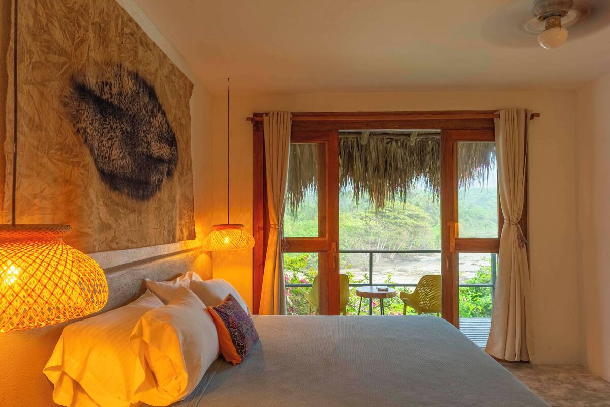 A bedroom with a large bed, a hanging light fixture, and a view of a balcony with outdoor seating.
