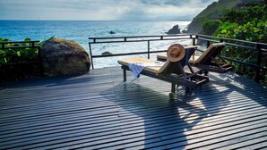 Terraza de madera con sillones de descanso y un sombrero, con vistas a una playa rocosa y un cielo despejado.