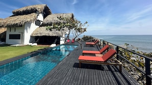 Un área de piscina con cubierta de madera, sillones de descanso y un edificio con techo de paja.