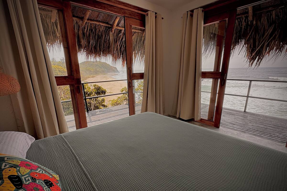 A room with a bed, a window with a view of the beach and ocean, and a thatched roof.