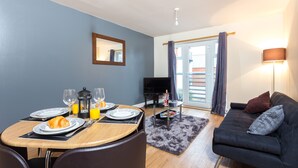 Un coin repas avec une table dressée pour deux personnes, un canapé, un téléviseur et un balcon avec des rideaux.