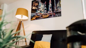 Un salon avec un canapé noir, un coussin jaune et gris, une lampe sur pied, une plante en pot et un tableau représentant un paysage urbain accroché au mur.