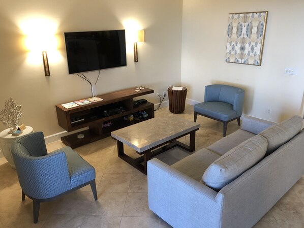 A living room with a flat-screen TV, two blue armchairs, a grey sofa, a coffee table, and a wooden entertainment unit.