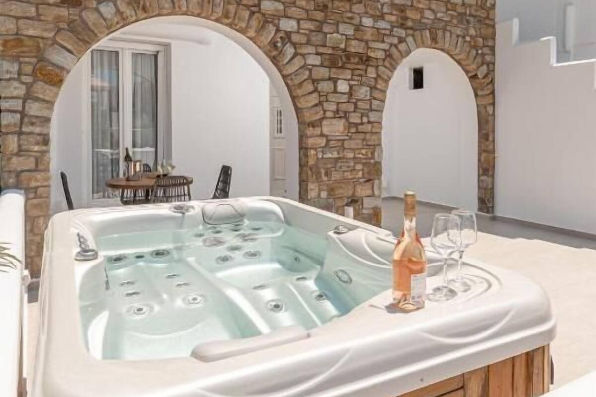 A modern outdoor hot tub area with stone arches, a wooden deck, and a table with a bottle and glass.