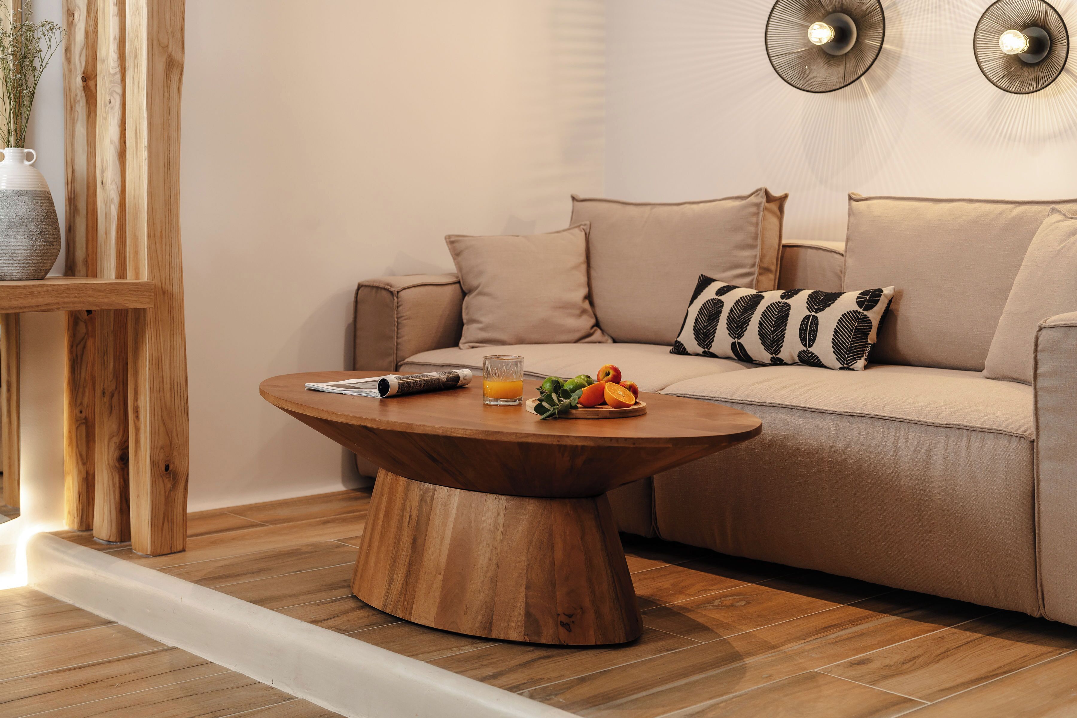 Un salon moderne avec une table basse ronde en bois, un canapé beige agrémenté de coussins à motifs et une petite table d’appoint sur laquelle se trouvent un vase et des fruits.