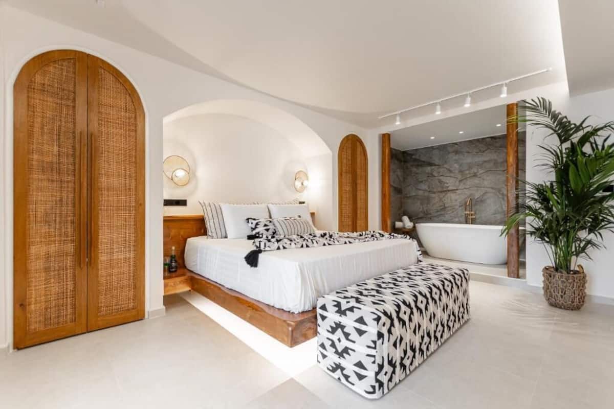 A modern bedroom with a large bed, a bench, a bathtub, and a potted plant.