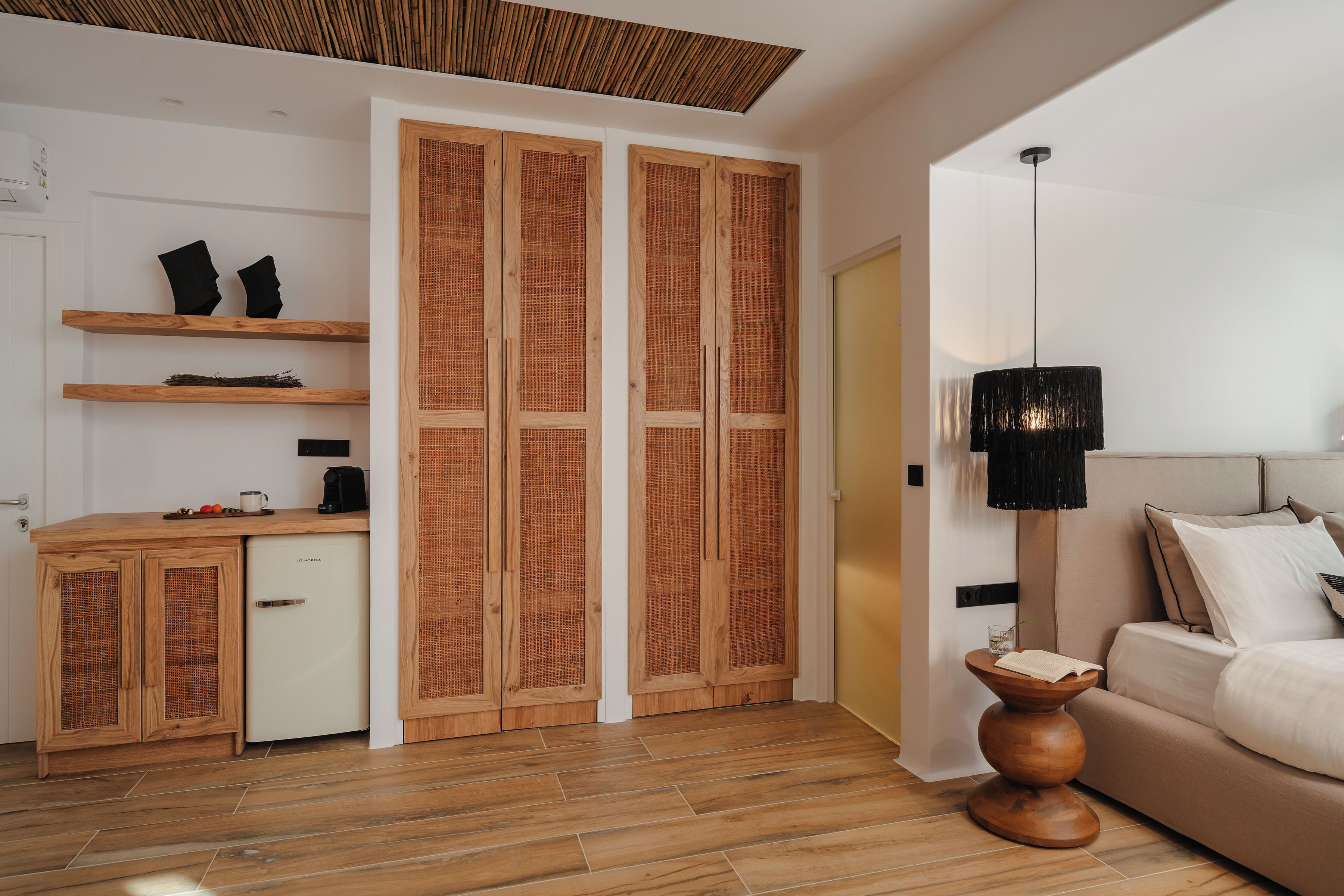 Un dormitorio moderno con suelo de madera, una cama con ropa de cama blanca, una mesita de noche con lámpara y un armario con panel trasero tejido.