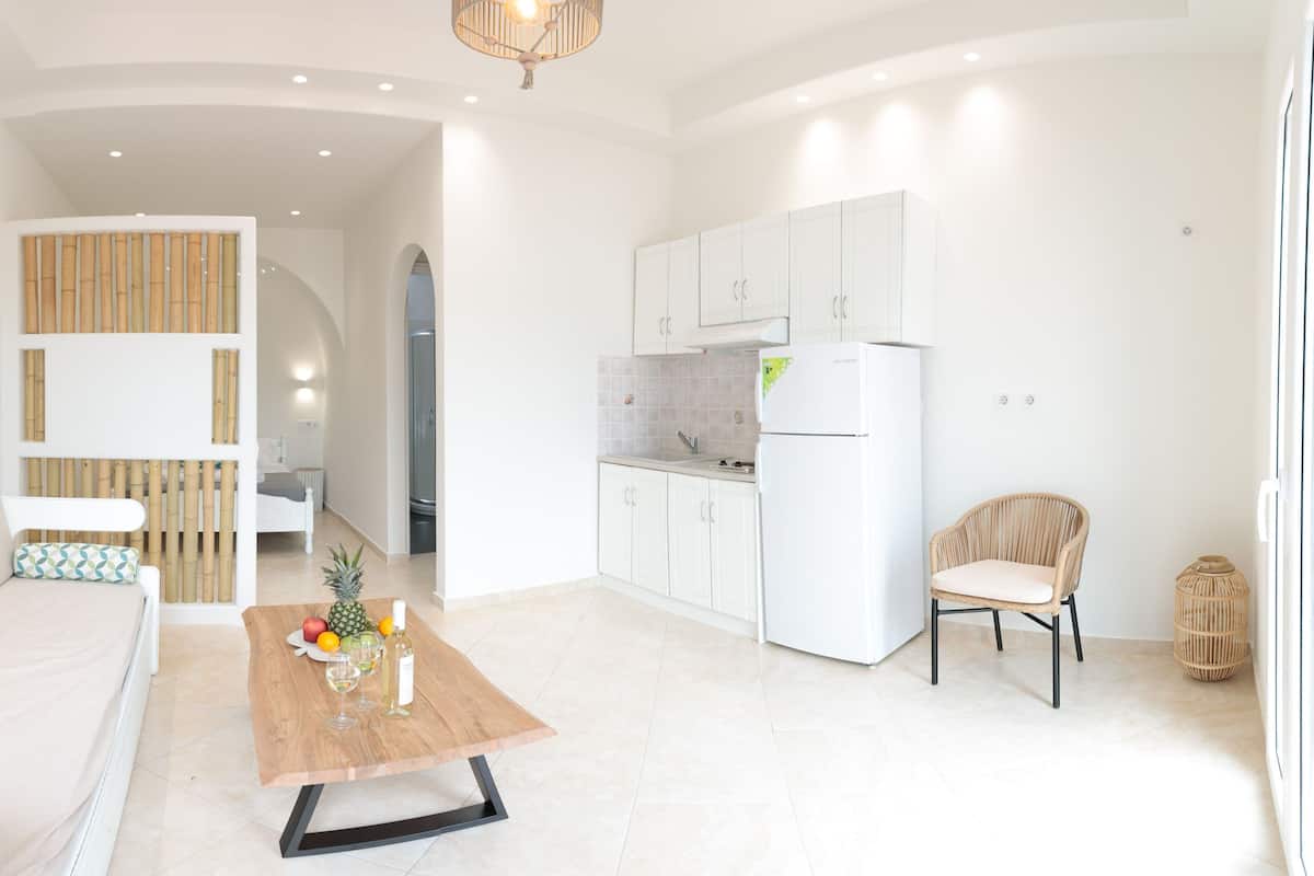 A modern living room with a sofa, a wooden coffee table with fruits and a wine glass, a wicker chair, and a kitchen area with white cabinets and a refrigerator.