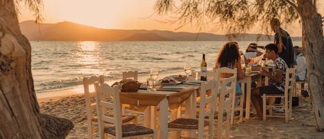 A beachside dining setup with tables and chairs, overlooking the sea and mountains.
