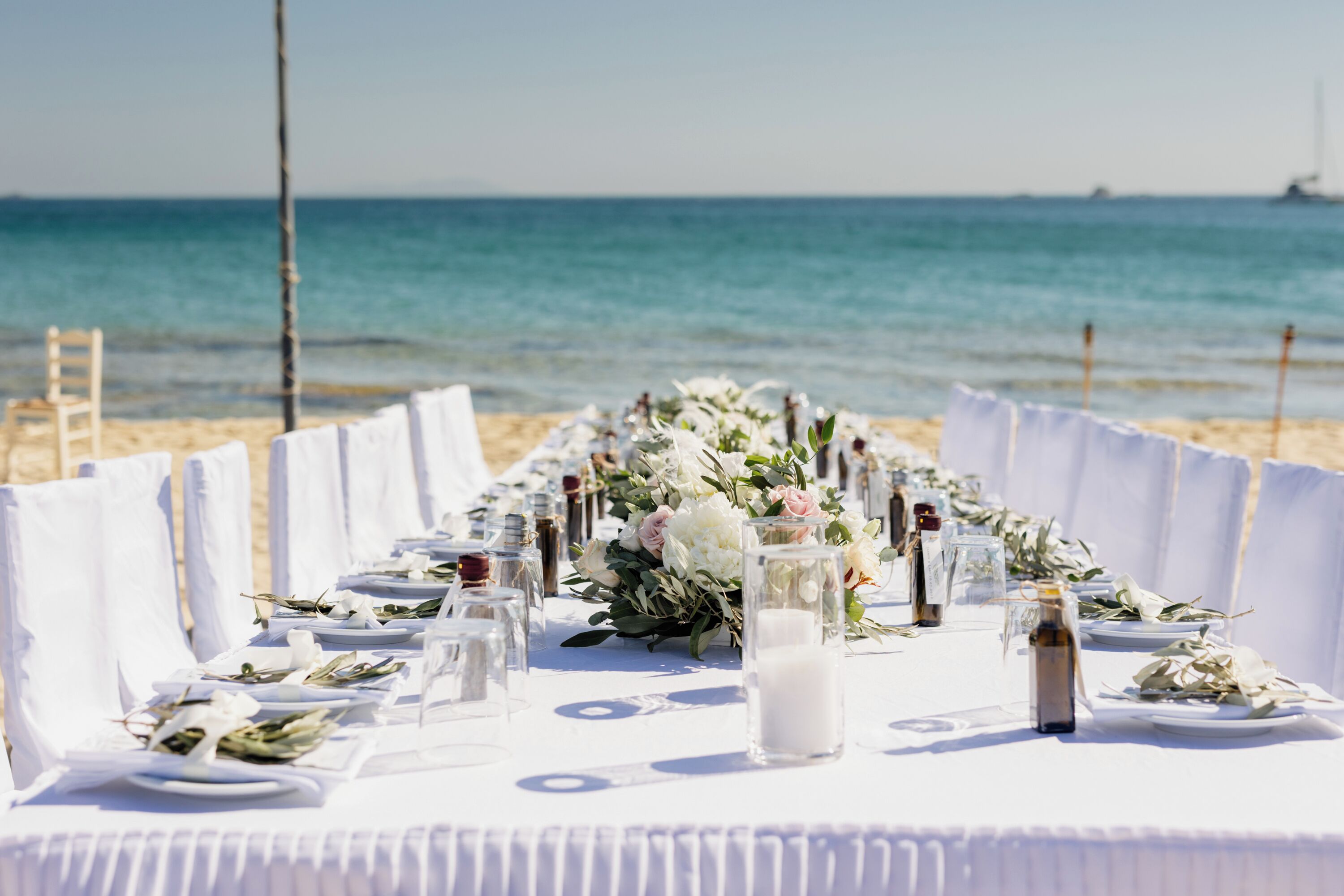 A long table set up on a beach with white tablecloths and floral centerpieces.