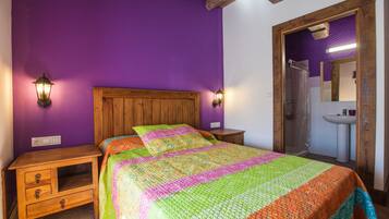 Quarto com cabeceira de madeira, colcha colorida, duas mesas de cabeceira e banheiro visível por uma porta aberta.