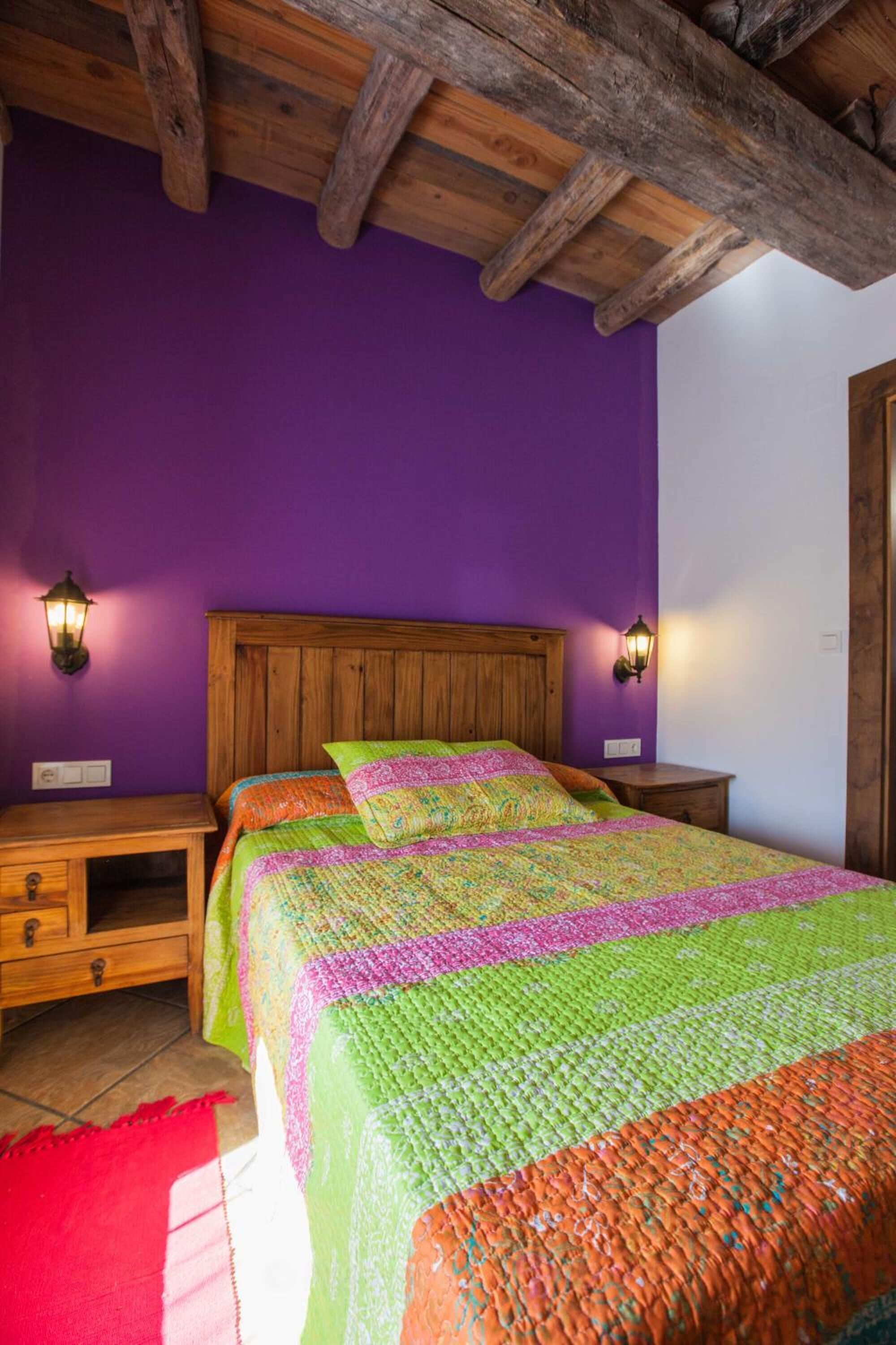 Quarto com cabeceira de madeira, paredes roxas e um edredom colorido.