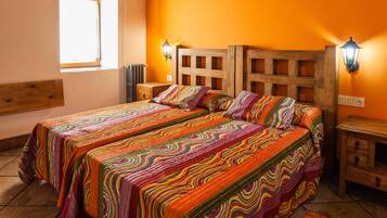 A room with a wooden ceiling, two beds with colorful bedding, a window with blinds, and a nightstand with a lamp.