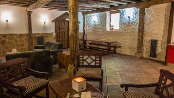 Un interior rústico con vigas de madera, paredes de piedra y una chimenea de piedra. Se observan mesas y sillas de madera, además de un banco de madera.