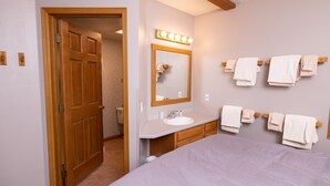 Um quarto com uma cama, um banheiro com pia e espelho, e uma porta que dá para outro cômodo.