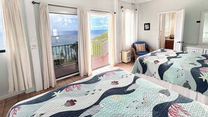 Une chambre avec deux lits, un ventilateur de plafond, un balcon avec des rideaux et une vue sur la mer.