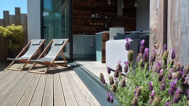 Eine Holzterrasse mit Liegestühlen, eine Glastür zu einer modernen Küche und ein Holzzaun.