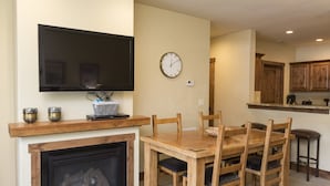 A living room with a fireplace, a TV mounted on the wall, and a dining area with wooden furniture. - Comfortable Condo - Convenient Upper Village Location (Whitefish)