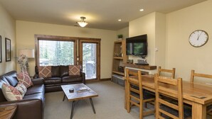 A living room with a sofa, coffee table, and wooden dining table with chairs. There is a television on a stand and a clock on the wall. - Comfortable Condo - Convenient Upper Village Location (Whitefish)